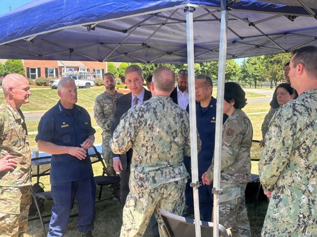 A group of both uniformed Air Force and Navy personnel stand around two men in suits. They are all standing outdoors under a pop-up canopy.